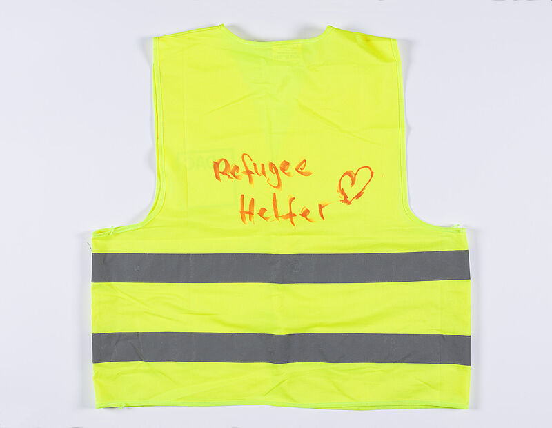 Gelbe Warnweste Größe 2XL, Vorderseite grauer Aufdruck "ADAC", Rückseite Handschrift in rot "Refugee Helfer (Herz-Symbol)", 2015