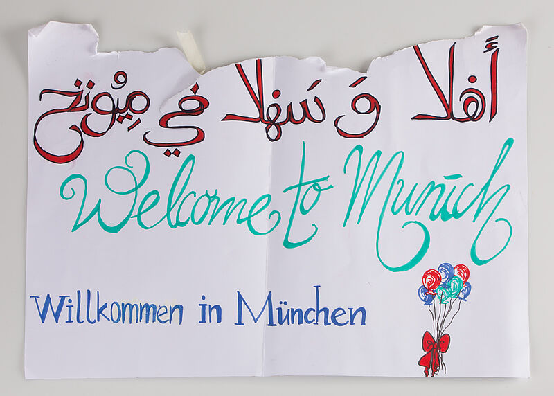 Plakat "Welcome to Munich Willkommen in München مرحبا بكم في ميونيخ", Handschrift rot-grün-blau auf weißem Hintergrund, 2015