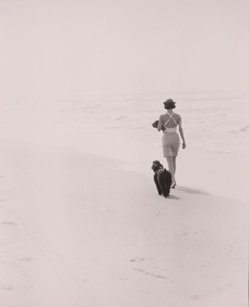 Hubs Flöter, Gaby Flöter Rückenansicht, spaziert am Strand mit Königspudel, kurzer Hose, Badeanzug und Hut